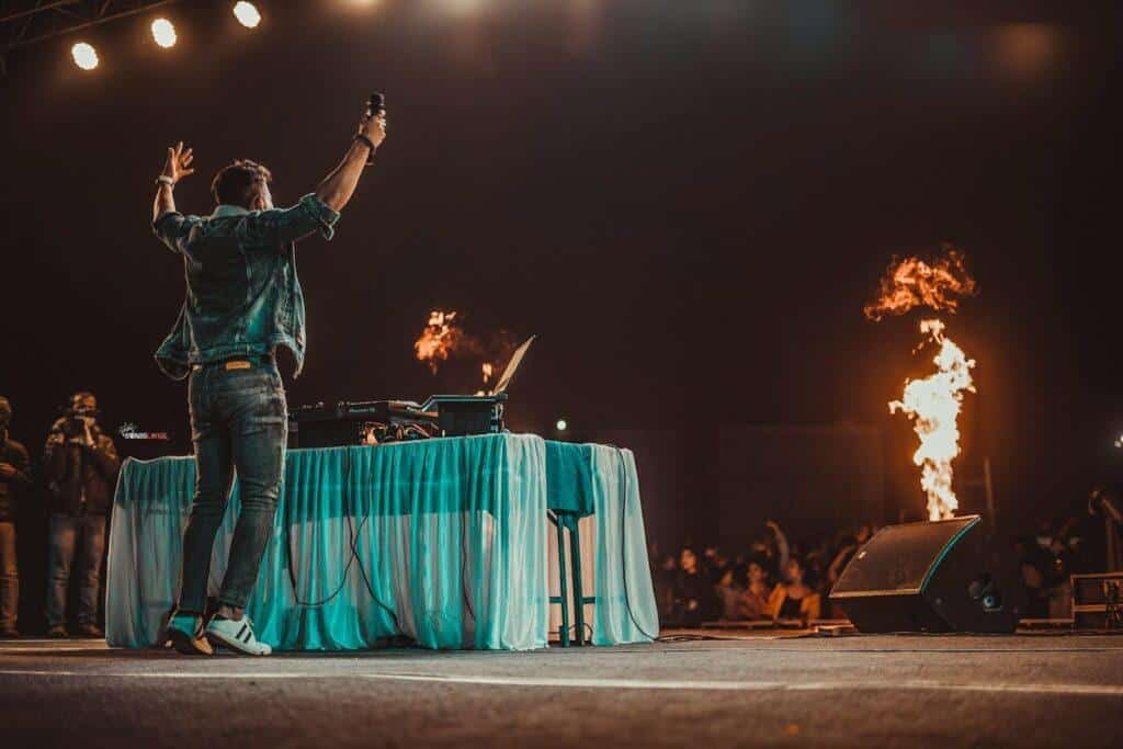 Alt: A DJ with their arms in the air, a pyrotechnic jet going off nearby