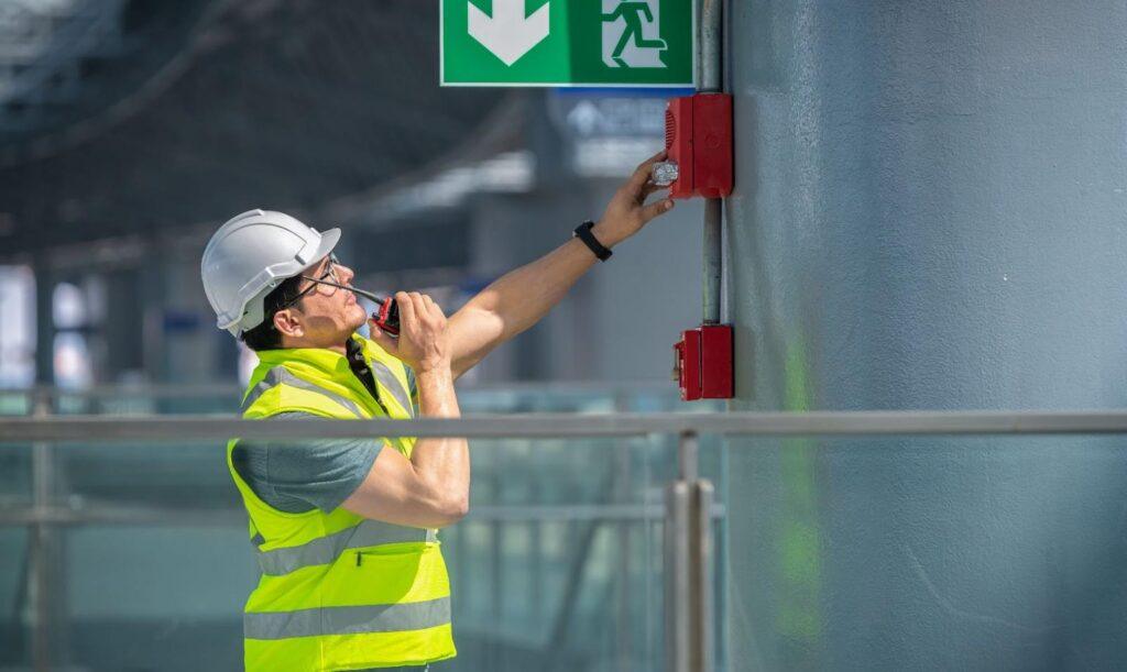 A fire watch guard wearing a safety uniform and a helmet checking the fire alarm emergency system in an industrial factory