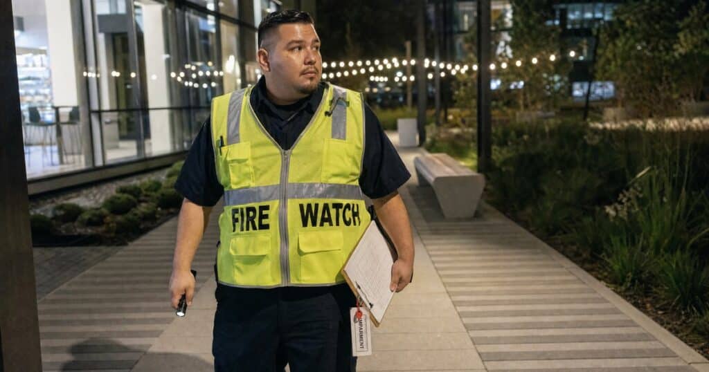 Fire watch guard on night patrol with a clipboard and hi-vis FIRE WATCH vest outside a San Jose office campus